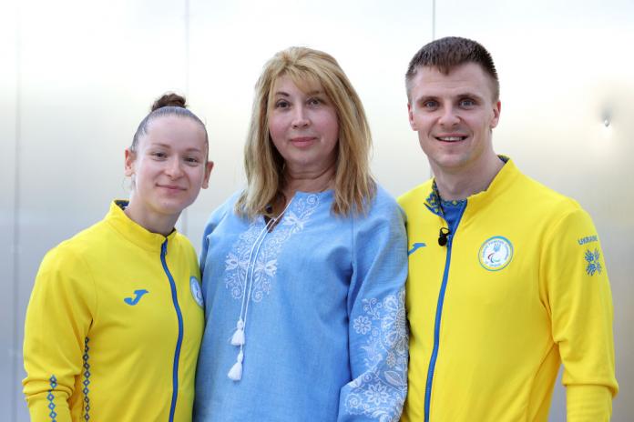 A woman and a man in yellow Team Ukraine jackets stand on either side of a woman in a blue shirt, with al three smiling for the camera