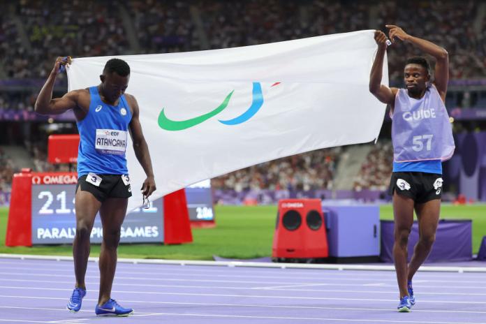 Two sprinters carry opposite sides of a white flag that has the Paralympic Agitos on it