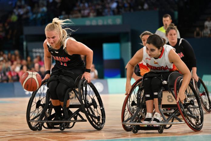 Two women, one in a black uniform and one in a white uniform, race after a basketball during a wheelchair basketball match