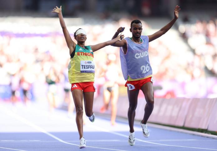 A woman wearing a blindfold and a man raise their arms in the air while running on a track