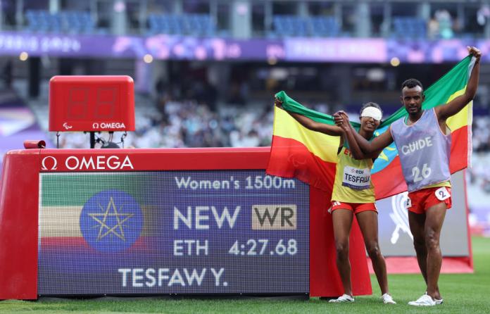 A runner wearing her blindfold and her guide celebrate a world record time with an Ethiopian flag next to their official time on a scorebaord