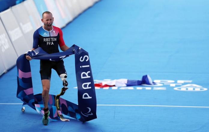Man in French traithlon one-piece screams in celebration, holding Paris 2024 finish line ribbon