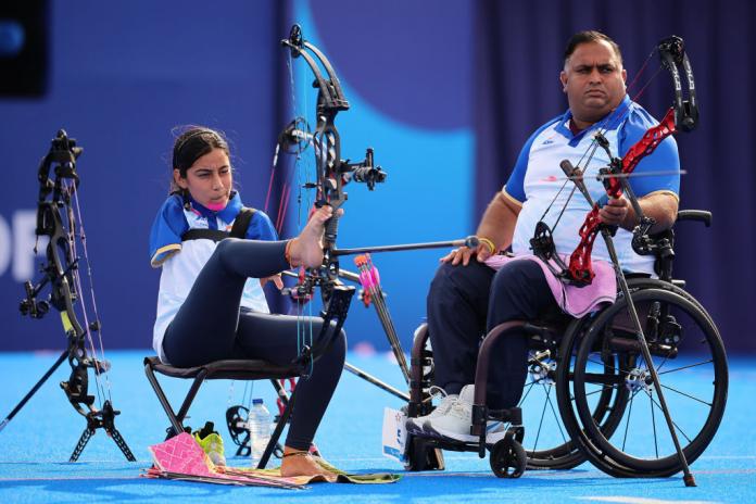 A woman uses her feet to aim an archery bow. Next to her, a man in a wheelchair holds a bow and looks at where she is aiming.