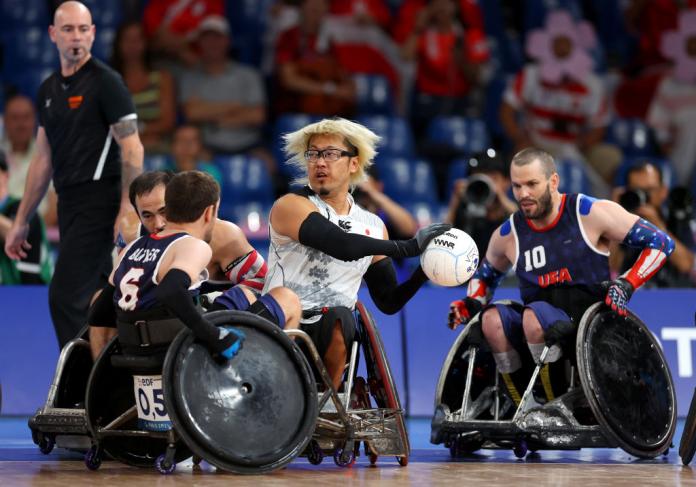 A man in a wheelchair in a white uniform holds a ball away from three other men in wheelchairs in blue uniforms