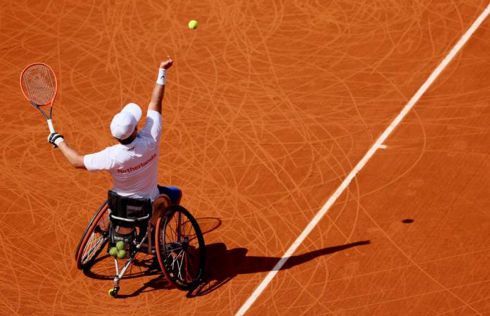 A man in a wheelchair throws a tennis ball in the air and prepares to hit it with his tennis racket