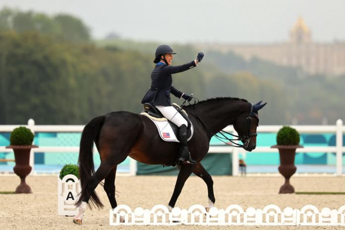 A woman rides a dark brown horse and lifts up an arm in celebration. A palace is in the background.