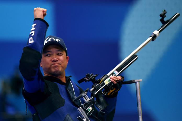 A man in a dark blue jacket and baseball hat lifts one arm in the air in celebration and holds his shooting Para sport rifle in his other hand.