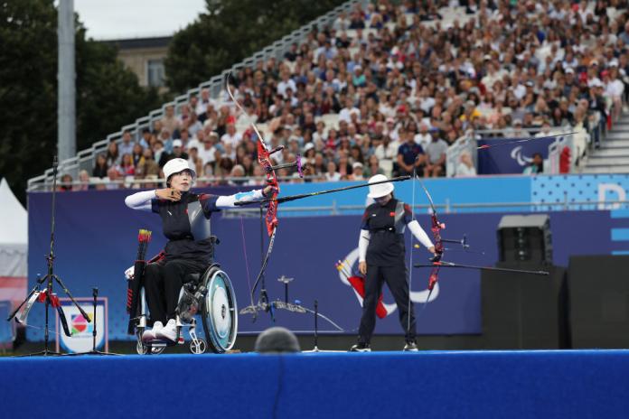A woman in a wheelchair aims a bow and arrow as a crowd watches on.