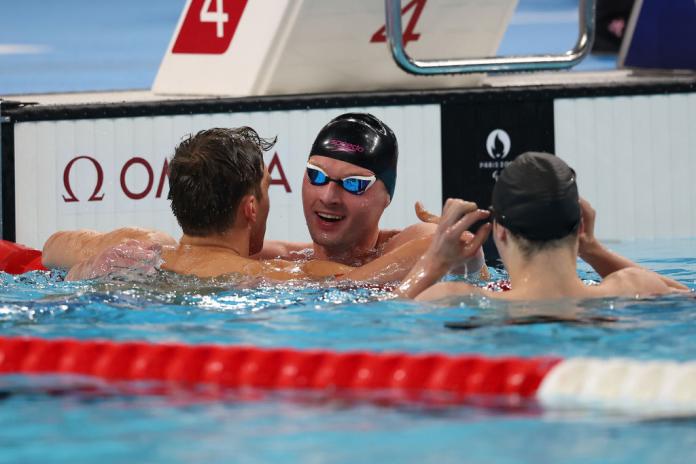 Three men are in a swimming pool, two are congratulating a third, who is wearing a swim cap and goggles.