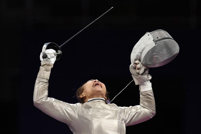 A woman competing in wheelchair fencing holds up her sabre in one hand and her face shield in the other and yells passionately