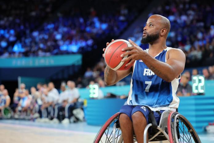 Un jugador de baloncesto en silla de ruedas con uniforme del equipo francés sostiene una pelota de baloncesto y la sostiene en preparación para disparar