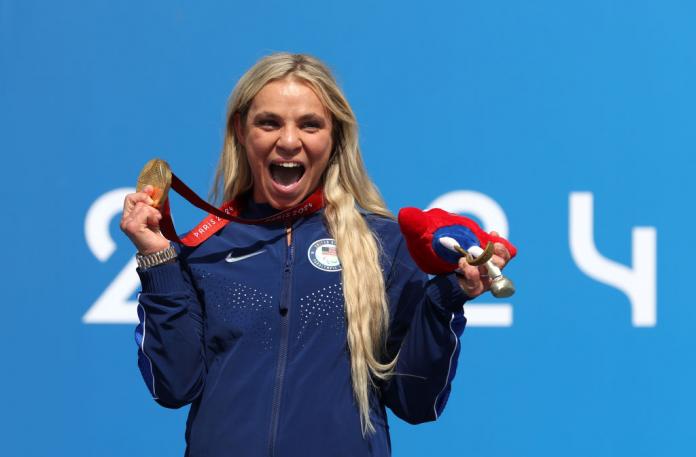 An American cyclist with long blonde hair in a blue jacket celebrates with a gold medal