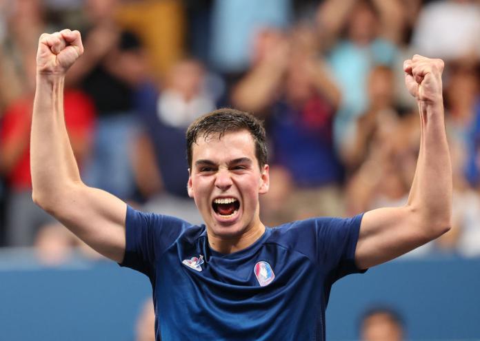 A man in a Team France shirt raises both arms in the air and yells in celebration