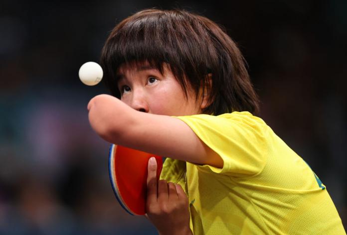 A table tennis player in a yellow shirt keeps her eye on the ball as she serves