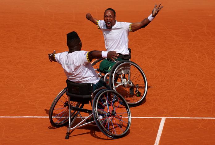 Two men in wheelchairs on a clay tennis court put their arms up in the air, one man yelling in celebration