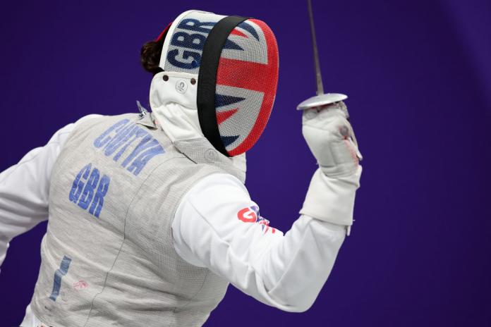 A person wears a wheelchair fencing uniform, and a face shield decorated with the union jack, and holds a sabre in one hand