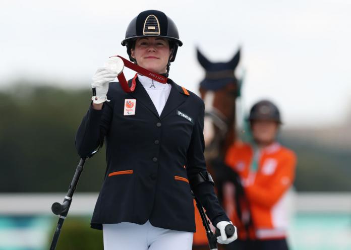 A woman stands in front of a horse and holds up a silver medal