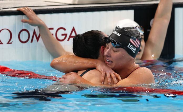 At the end of a swimming pool, a woman in a black swim cap with a USA flag on it hugs another swimmer