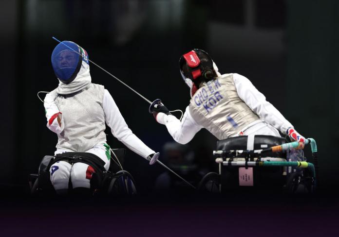 Two wheelchair fencers face with the Korean leaning forward to strike Italy whose face we see react through the blue mask