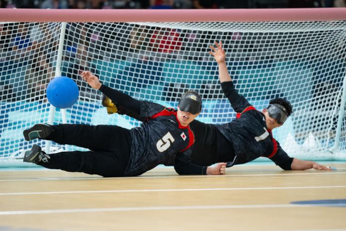 Two goalball athlete dive in front of their net to stop a blue ball