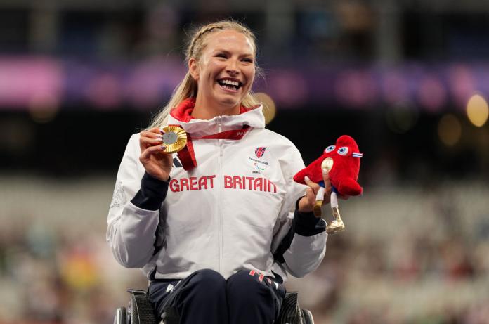 A woman in a wheelchair holds a gold medal and a Paris 2024 mascot and smiles