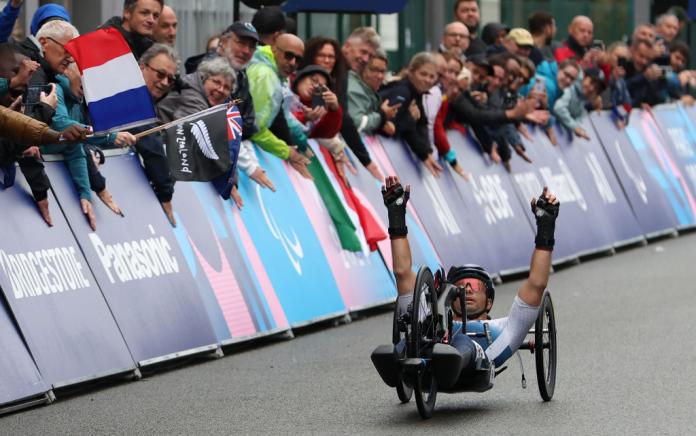 A man on a handcycle raises his arms up in the air in celebration, as a crowd of people watch on the side of the road