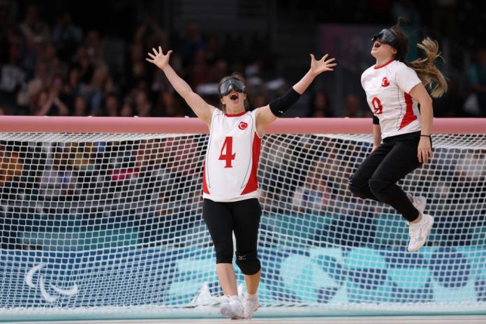 Two women in Turkiye uniforms and black eye shades celebrate winning a goalball game, one woman raises her arms in the air and the other jumps off the ground