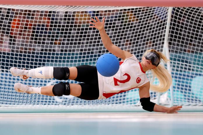 A woman plays goalball in a Turkiye uniform and black eyeshades, diving across a net to block a blue ball