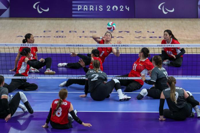 Dos equipos juegan voleibol sentado, uno con camisetas rojas y otro con camisetas grises y negras. Una jugadora vestida de rojo levanta las manos para jugar al voleibol, que está en el aire.