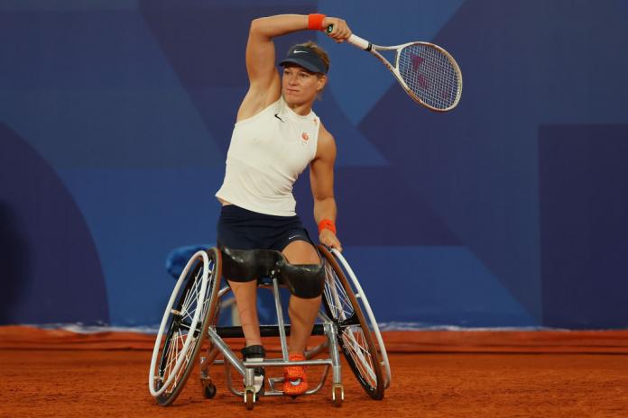 A woman in a wheelchair plays tennis, and has her racket lifted in the air after hitting the ball