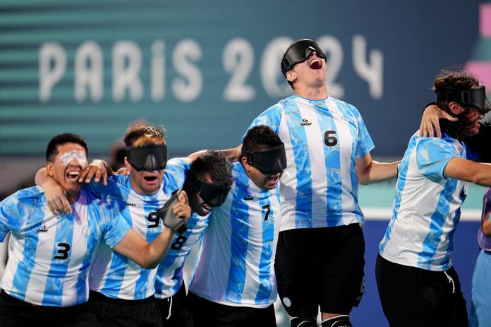 Hombres con camisetas azules y blancas del fútbol argentino con gafas negras se paran en fila con los brazos alrededor de los demás, gritando en celebración