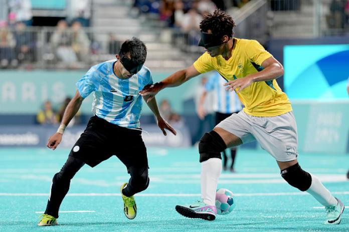 Un hombre con una camiseta amarilla de fútbol brasileño y un anteojo negro juega una pelota de fútbol con el pie, mientras que un hombre con una camiseta azul y blanca de fútbol argentino, también con un antifaz, corre hacia él