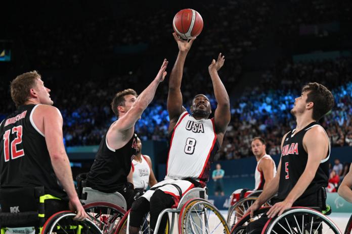 Un hombre con un uniforme de baloncesto en silla de ruedas de EE. UU. alcanza una pelota de baloncesto, con jugadores con camisetas de Canadá rodeándolo