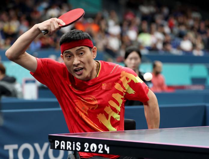 A man in a red team China shirt and red headband plays the ball in a Para table tennis match