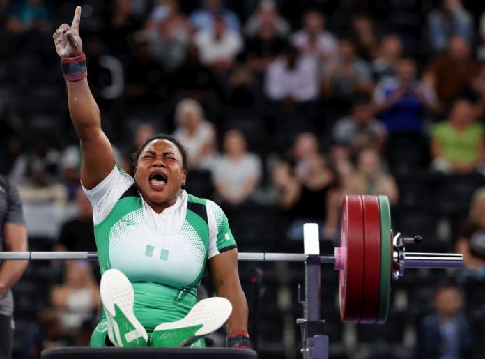 Un Para levantador de pesas con uniforme del equipo de Nigeria se sienta en un banco de levantamiento de pesas y levanta un brazo en el brazo, gritando en celebración