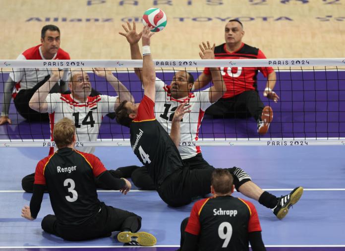 Two teams play sitting volleyball. A player on one side of the net strikes the ball, while two players on the other side of the net put their arms up to block it.