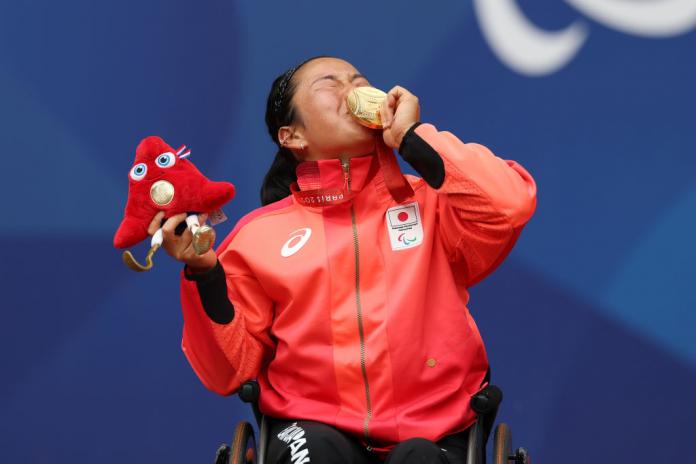 A woman in a wheelchair wearing a Team Japan jacket kisses a gold medal while also holding the Paris 2024 mascot