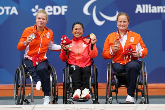 Three women in wheelchairs are on a podium. Two Dutch athletes hold up silver and bronze medals, while a Japanese athlete holds up a gold medal