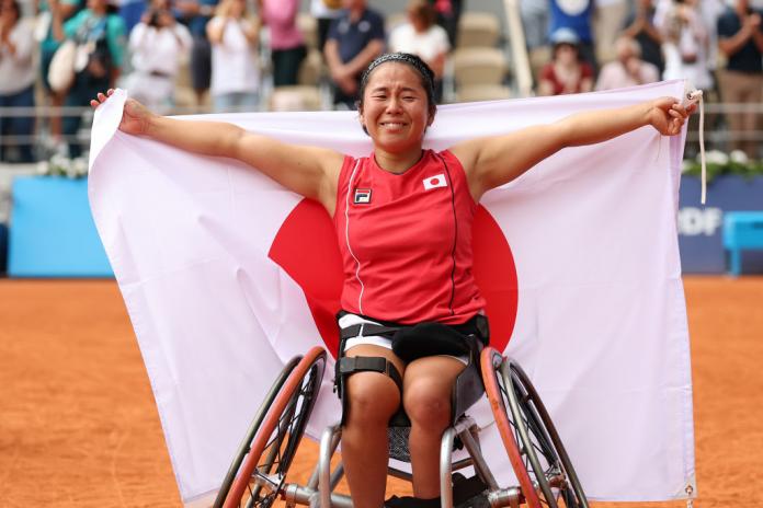 A woman in a wheelchair poses on a wheelchair tennis court, holding up a Japanese flag behind her shoulders