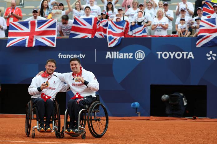 Two wheelchair tennis players in Great Britain jackets hold up gold medals in front of a crowd of people waving Great Britain flags
