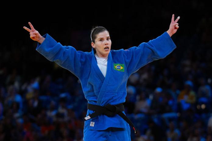 A woman in a blue judoka uniform raises both arms in the air, making peace signs with her fingers
