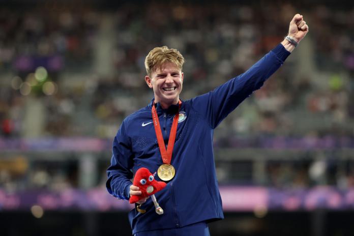 A man in a Team USA jacket wears a gold medal around his neck, holds the Paris 2024 mascot in one hand and raises his other hand in the air, smiling in celebration