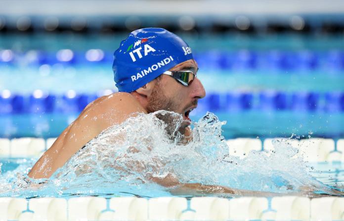 A Para swimmer in a blue Team Italy swim cap and goggles comes up out of the water to take a breath