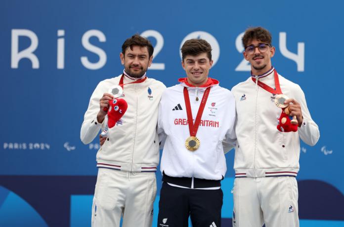 Three male cyclists, all dressed in white tracksuits, celebrate their medal wins on the podium