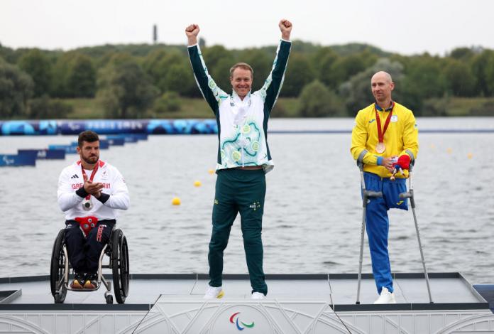 An Australian man in a white tracksuit top and green trousers lifts both hands above his year on the podium