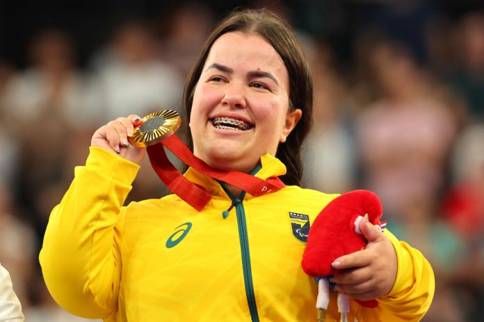 A woman in a Team Brazil jacket holds up a gold medal in one hand and carries the Paris 2024 mascot in her other hand