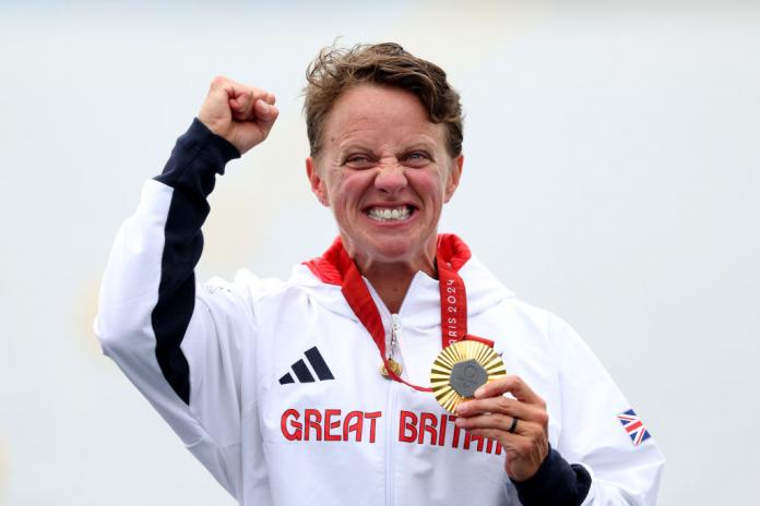 A female kayaker in a white tracksuit top punches the air with one hand and has a gold medal in the other 