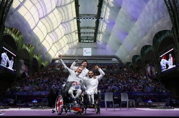 Three wheelchair fencing athletes pose for a photo in front of a full grandstand in Paris' Grand Palais, raising their arms in the air in celebration