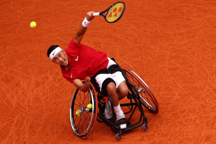 A man in a red Team Japan shirt plays wheelchair tennis, and looks at the tennis ball in the air that he just hit