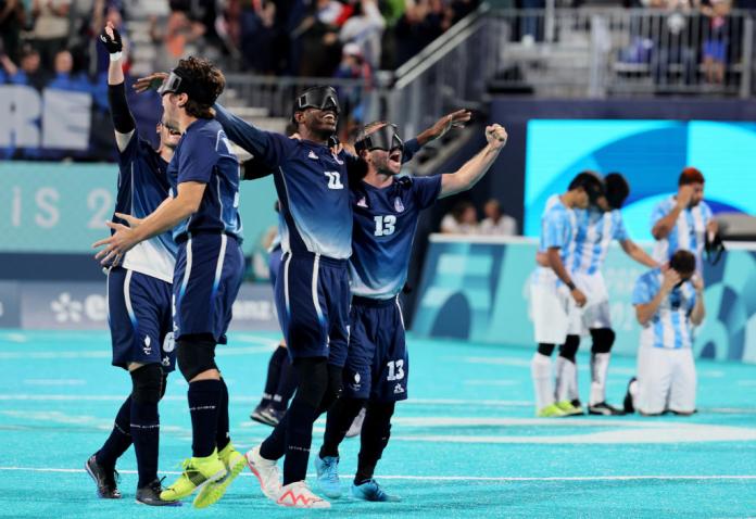 A group of French blind football players in blue uniforms and black eyeshades raise their arms in the air in celebration